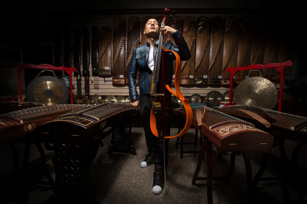 Dana Leong with electric cello in a hall of traditional Asian instruments, gongs and zithers behind him