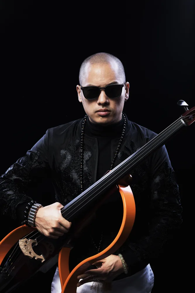 Studio portrait of Dana Leong in black against a black background, holding his electric cello, wearing sunglasses