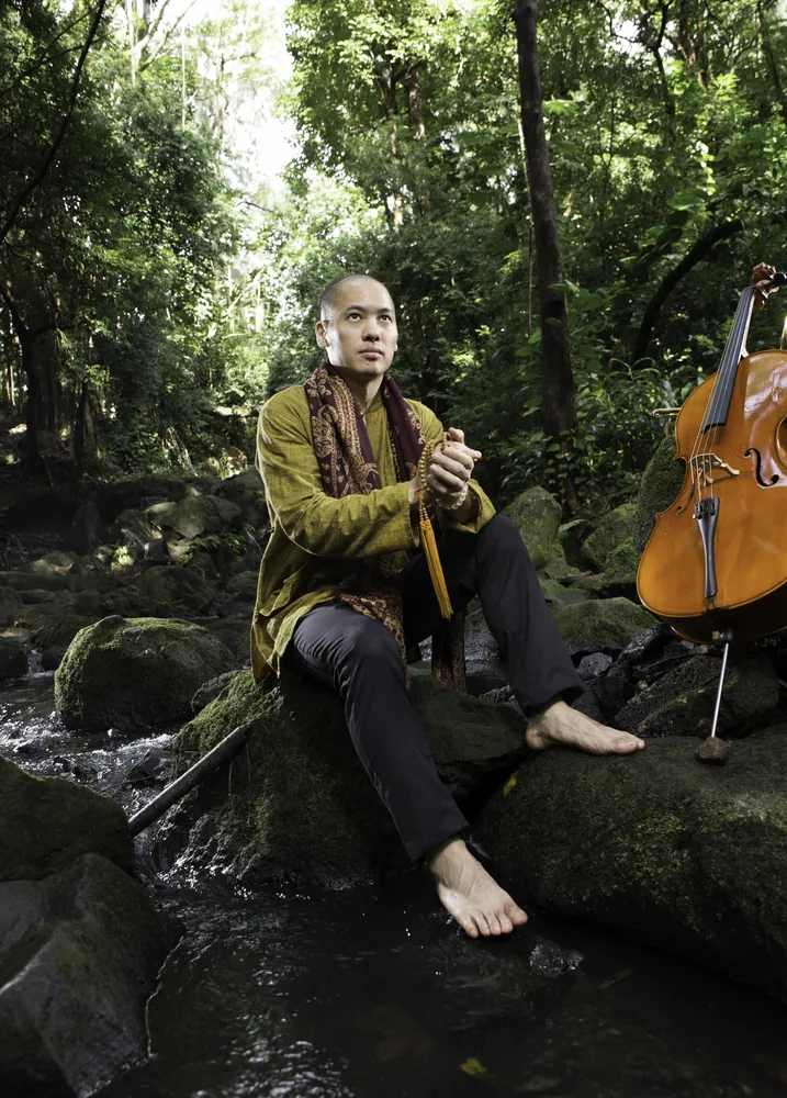 Dana Leong seated barefoot on mossy rocks in a rainforest stream with his acoustic cello beside him